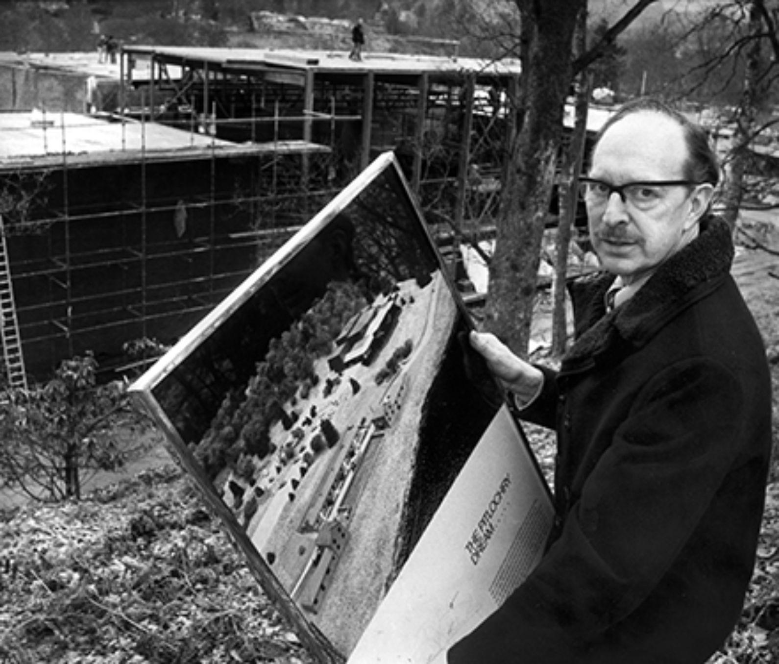 A black and white image of a man holding a picture of what the construction in front of him will turn out to look like, in the background is scaffolding and foundations of the theatre