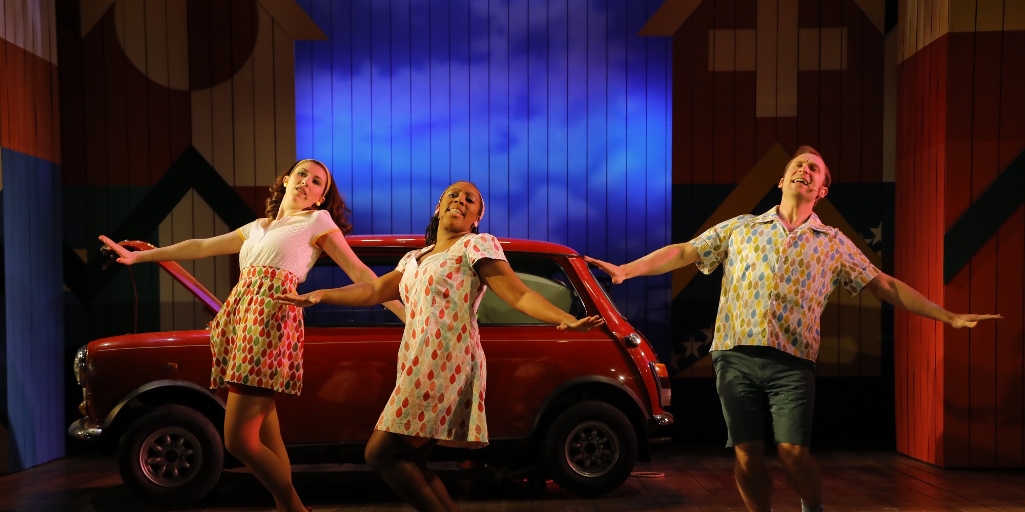 Three actors on stage all in a unified dance pose leaning back left with arms spread at their side, they are all standing in front of a red mini cooper with a blue sky design in the background