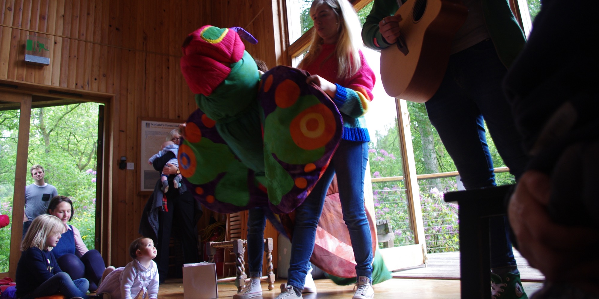 People inside a building made of wood, a person holds up a fabric made butterfly of various colours