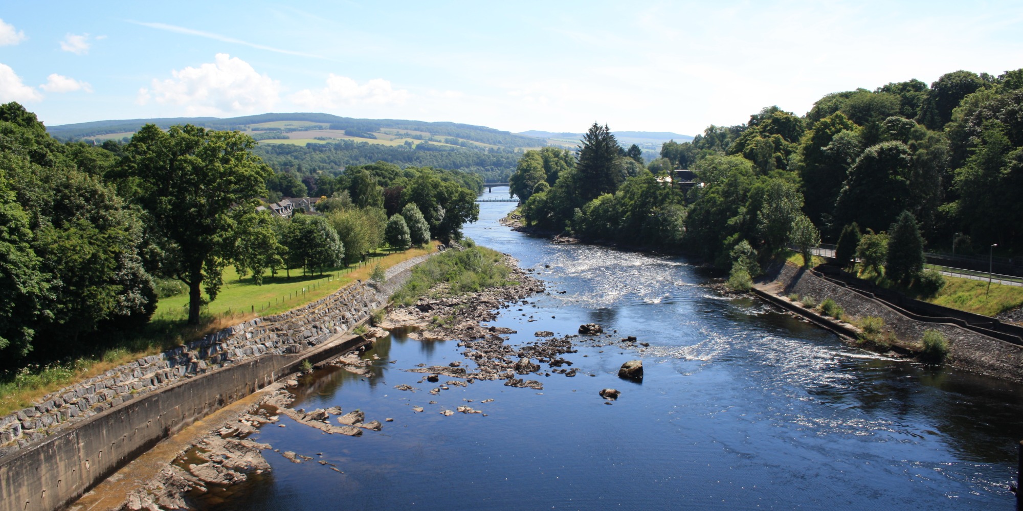 Image of a river with banks with trees on either side, in the distance is a bridge going over the river with blue skies and minimal clouds above