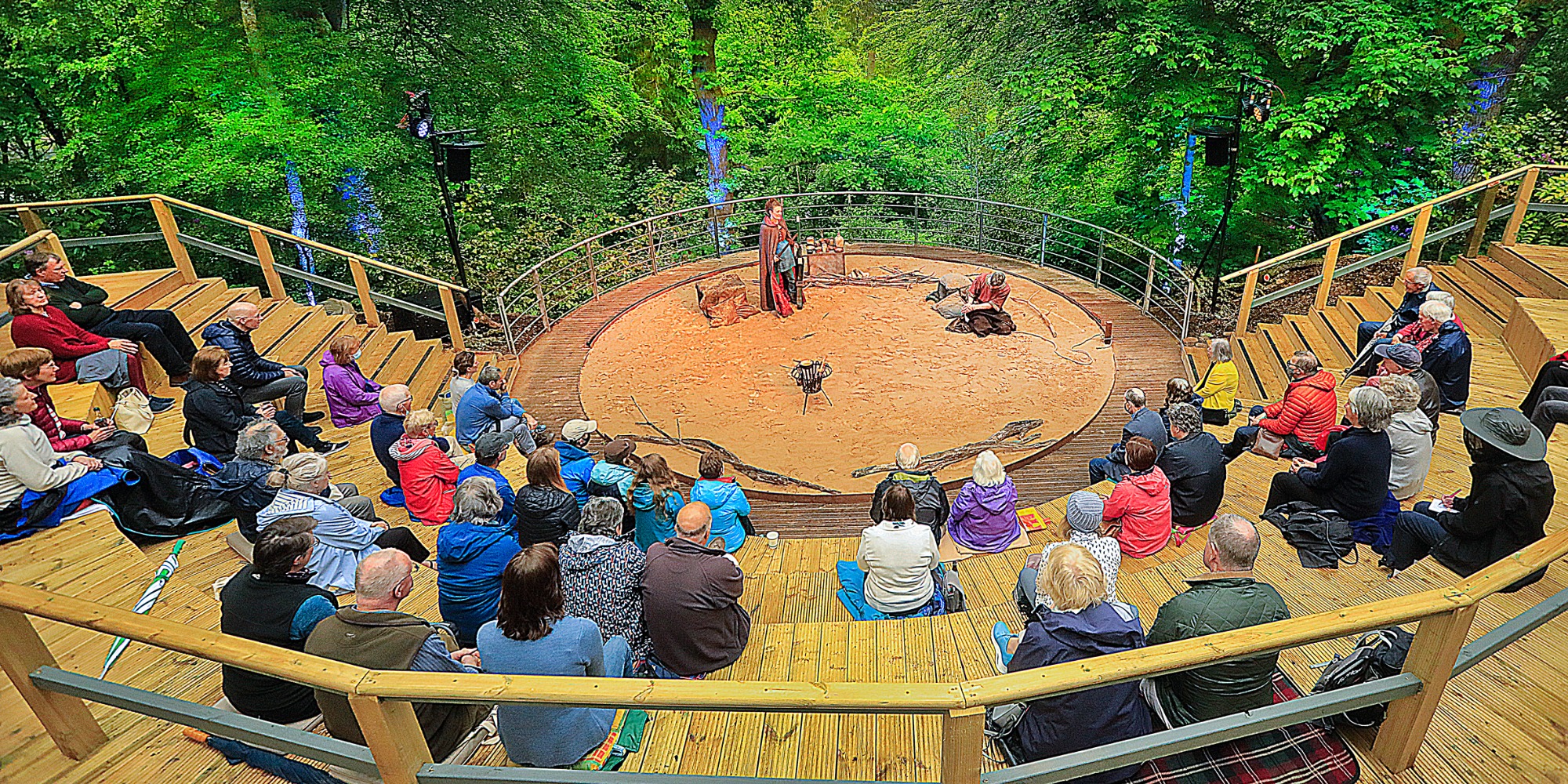 Image of the outdoor amphitheatre, wooden seating is located in a semi circle around half of the amphitheatre and on the staging area is an actor performing, a metal railing circles the outer rim and behind is a drop that leads in a garden with many green trees surrounding the top half of the image