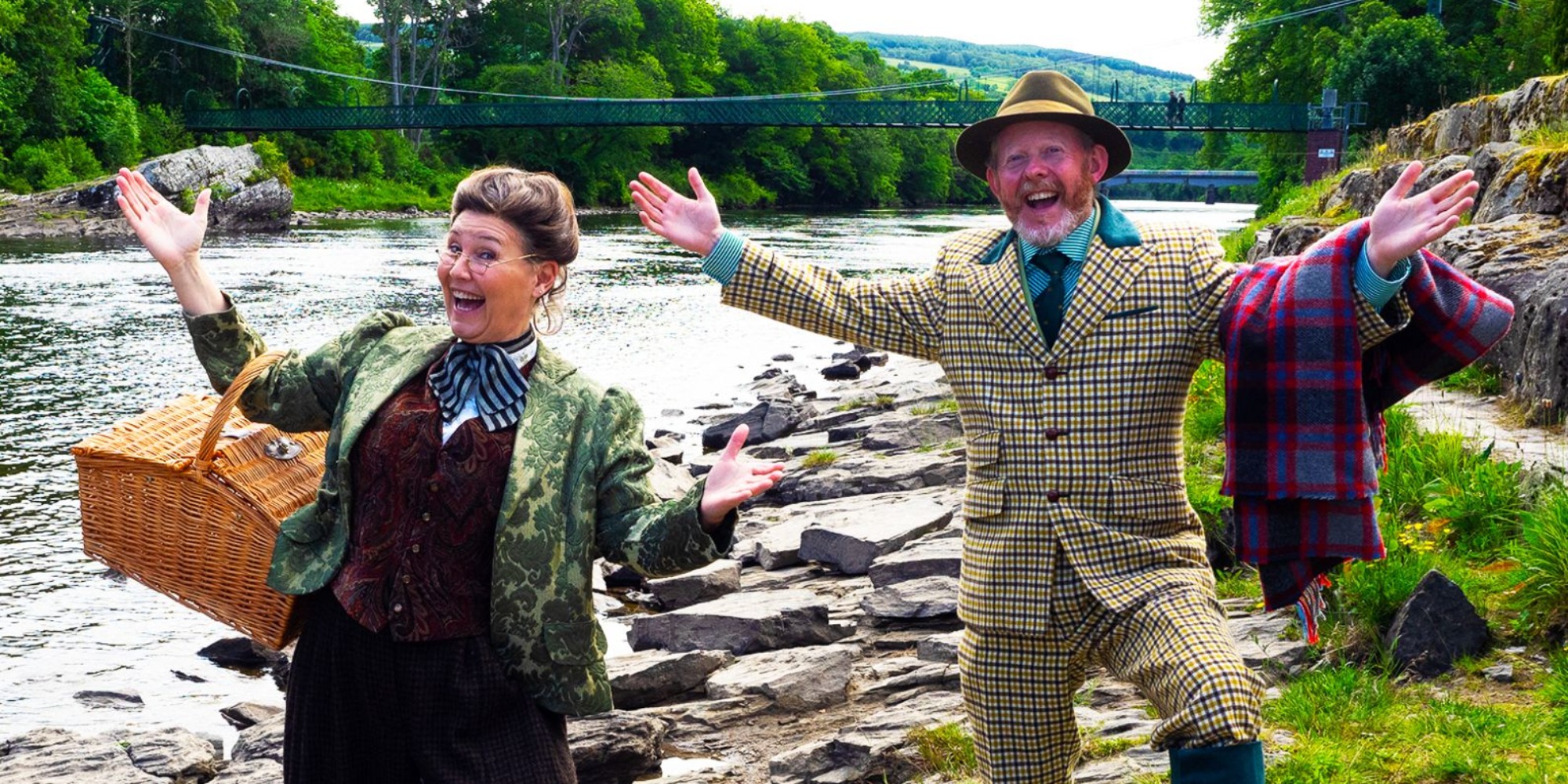 Two actors standing on the bank of the River Tummel, one dressed in a Tweed jacket and trousers with a red tartan blanket over their arm and the other in a floral jacket and a hamper over their arm, behind them is a green bridge going over the River
