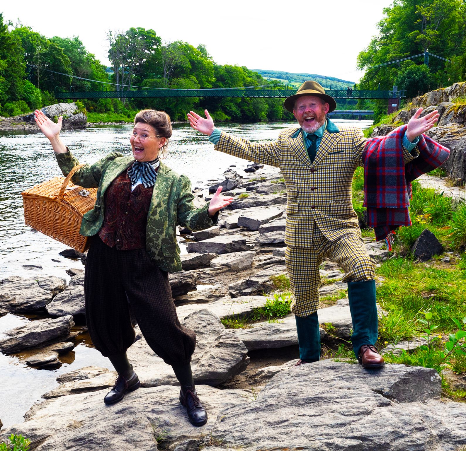 Two actors standing on the bank of the River Tummel, one dressed in a Tweed jacket and trousers with a red tartan blanket over their arm and the other in a floral jacket and a hamper over their arm, behind them is a green bridge going over the River