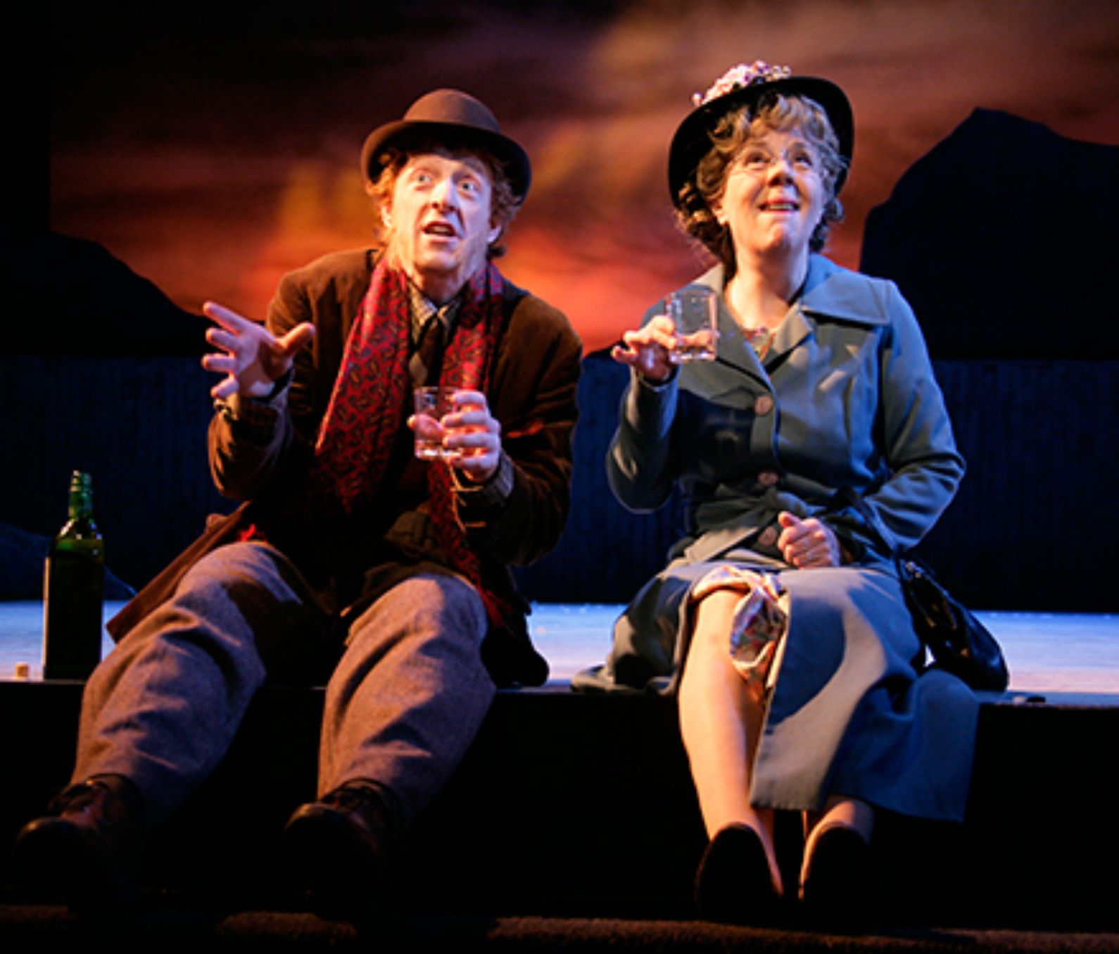 Two actors sitting on a stage looking into the audience both with looks of happiness on their face, both holding a glass with the actor on the right sitting beside a green bottle, the background is made to look like a red sky in the evening. Both are wearing hats and dressed for an outdoors setting with the actor on the left having a red scarf