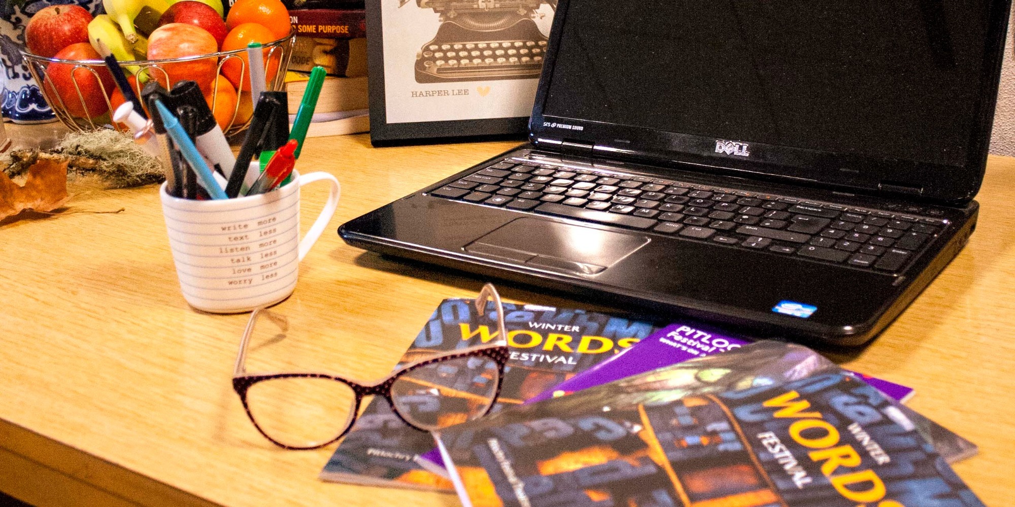 A laptop on a desk with flyers and a pair of glasses, on the left side of the desk is a cup full of pens and behind that is a fruit bowl