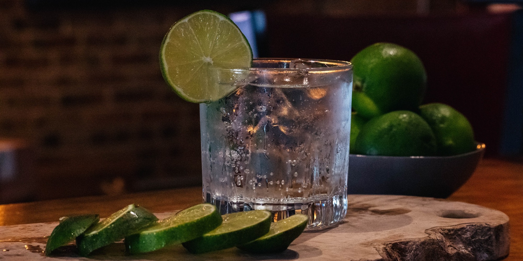 A glass of alcohol with condensation dripping down the outside of the glass with a slice of lime on the rim and ice cubes within. In the foreground is a sliced lime on its side and a bowl of limes behind the glass. The glass is on top of a sliced piece of wood.