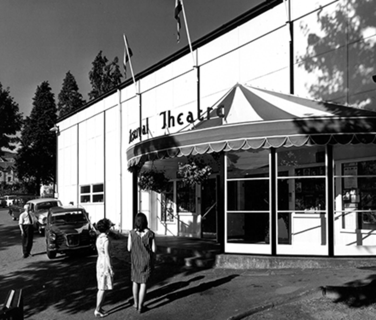 Black and white image of the entrance to the theatre with a canopy over the doors and the words 'Festival Theatre' written above, three people are approaching the door with two cars parked nearby. Flags have been arranged on poles above the canopy and stretch the guttering of the building