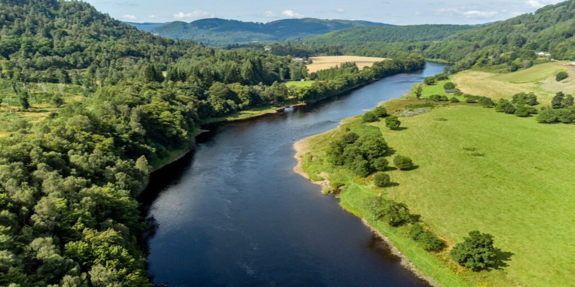 A large river running through the middle of the image with trees on the left hand side of the image and the background, on the right is a field with minimal shrubbery.