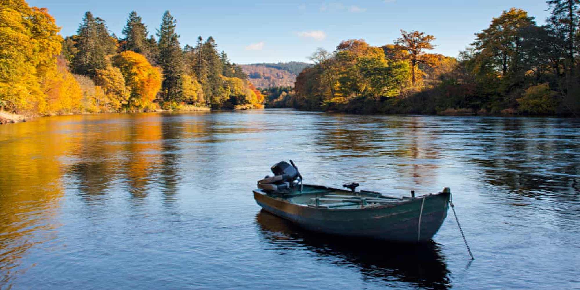A motorboat on in the foreground of the image on a body of water, in the background are trees in autumnal colours with blue skies above and minimal clouds.