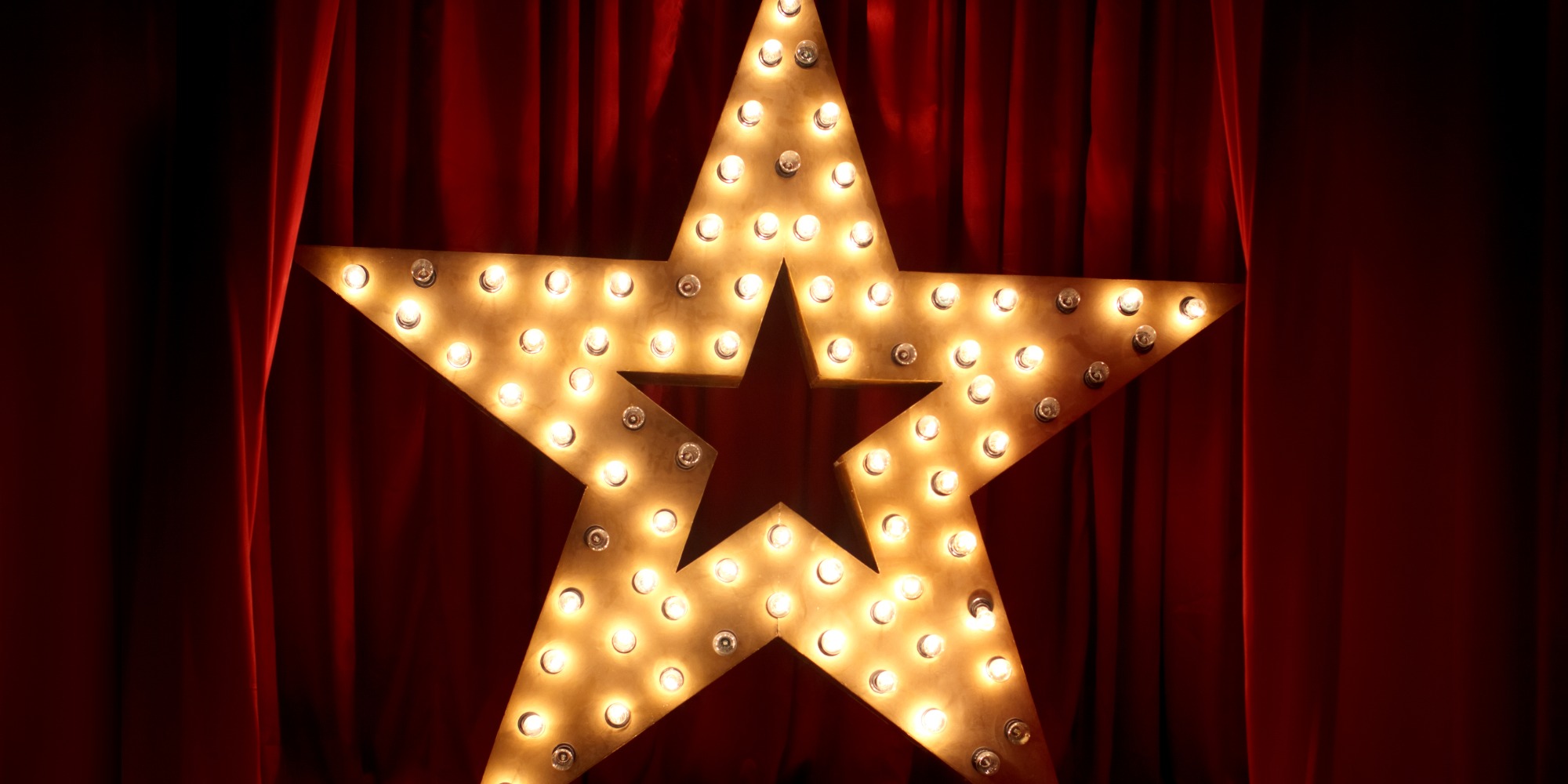 A five pointed star made up of a metal frame and many lightbulbs in front of a red Theatre curtain