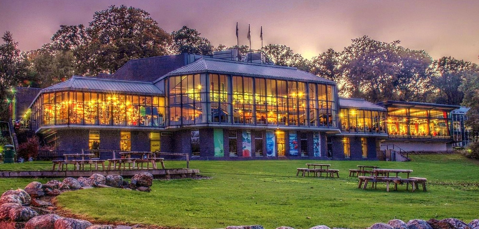 An image of the front of the main building of Pitlochry Festival Theatre with a full glass front and lights shining through each one, the ground outside is all covered in grass with wooden benches populating different parts and an evening sky above