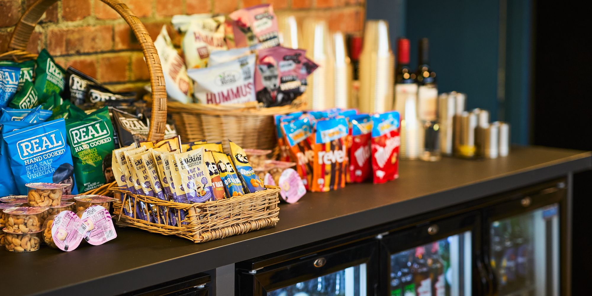 An image of a counter decorated with a variety of different chocolates, nuts and crips as well as cups on the right hand side of the image and fridges below the counter.