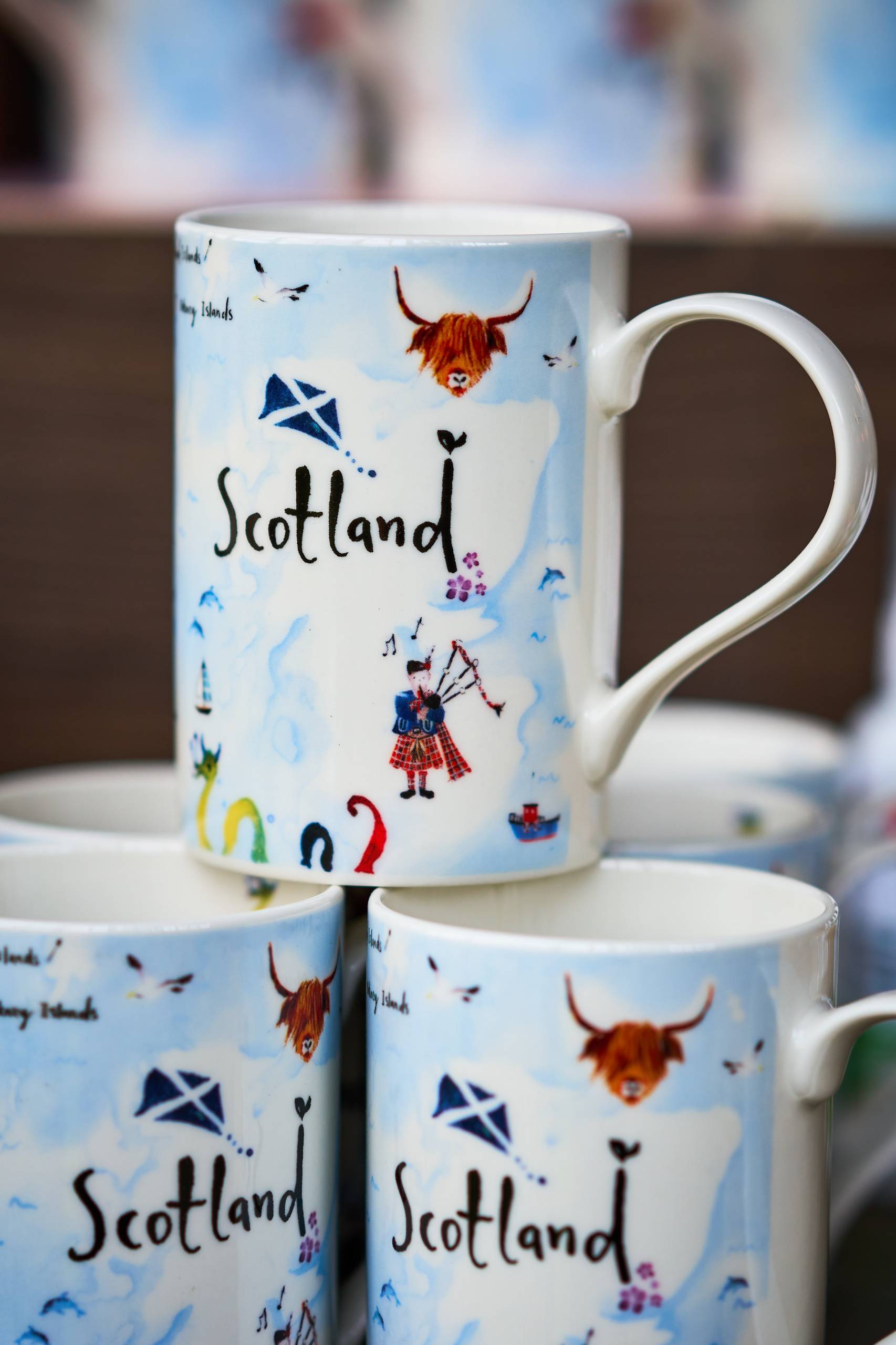 A coffee mug with the word 'Scotland' across it, the mug has a highland cow, someone playing the bagpipes and the loch ness monster on it