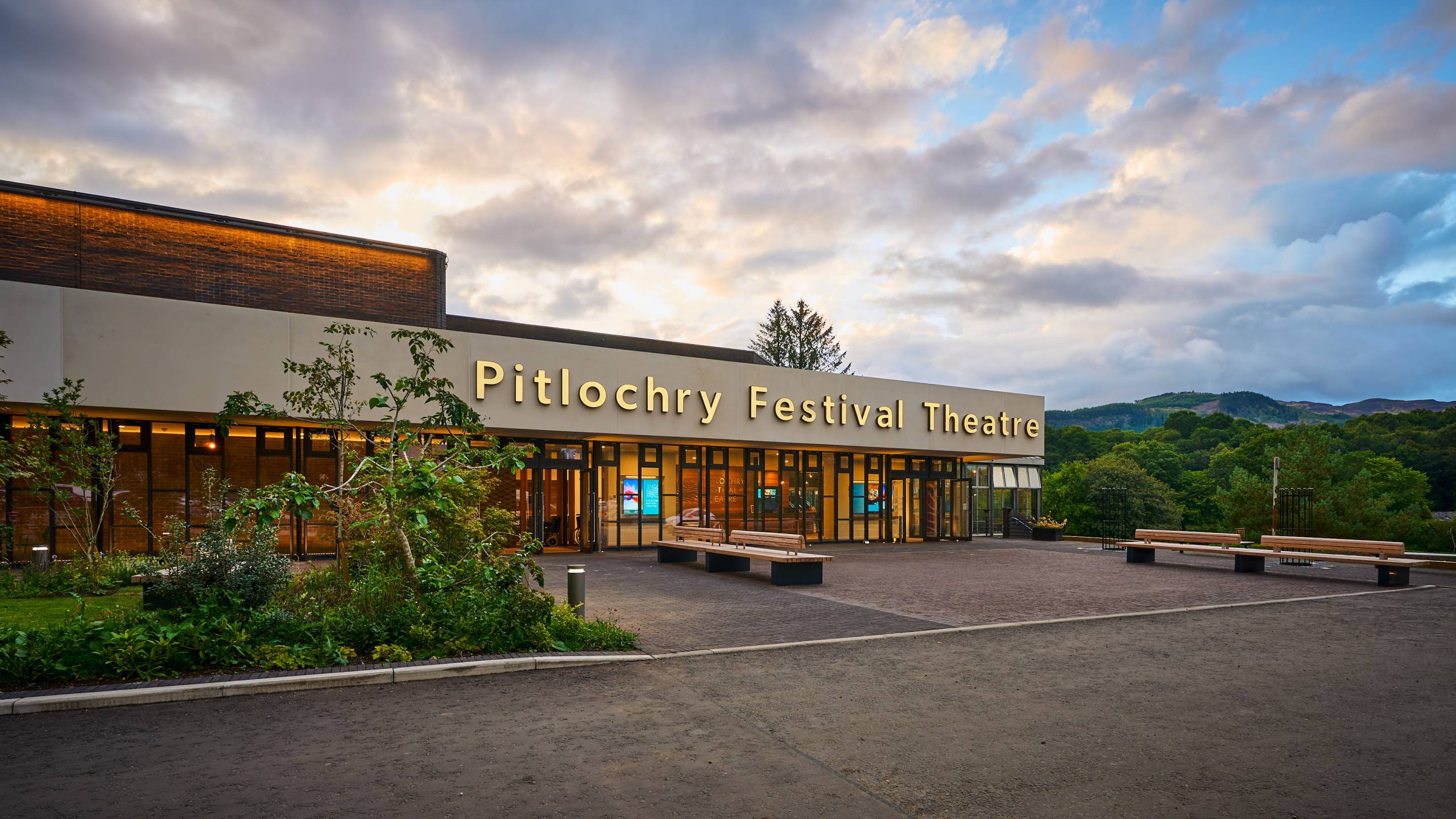 An image with a sign on the theatre's front entrance that says Pitlochry Festival Theatre with the sky and trees behind it
